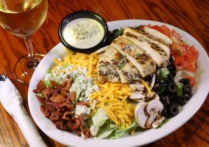 A salad topped with grilled chicken, bacon bits, bleu cheese, cheddar cheese, mushrooms, sliced olives and tomatoes, bleu cheese dressing and a glass of white wine