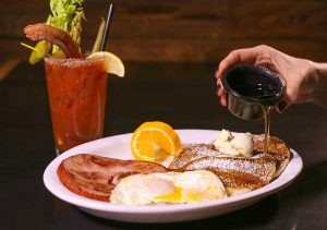 A Bloody Mary with a salted rim, Crispy bacon and veggie fixings sits behind a plate of ham, fried egg, orange slice and buttered pancakes topped with powdered sugar. A hand drizzles maple syrup onto the pancakes