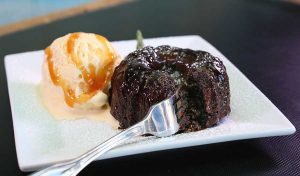 A fork cuts into a glistening chocolate lava cake plated with caramel drizzled ice cream and topped with powdered sugar