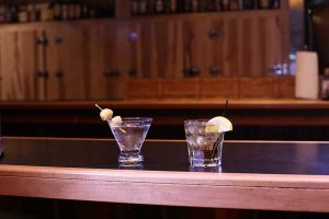 Two cocktails sit on the bar at Sam's Place, one with a straw and lemon, and another with two olives, straight