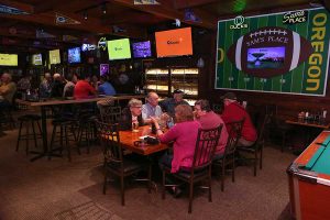 A group of Sam's Place patrons sits at the table below the Oregon football field and football framed television next to the pool table